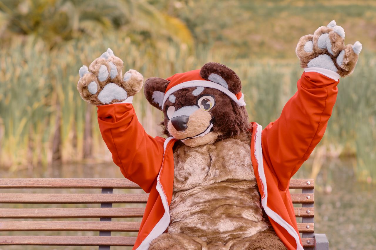 A furry character dressed in a Santa outfit sitting on a bench with arms raised outdoors.