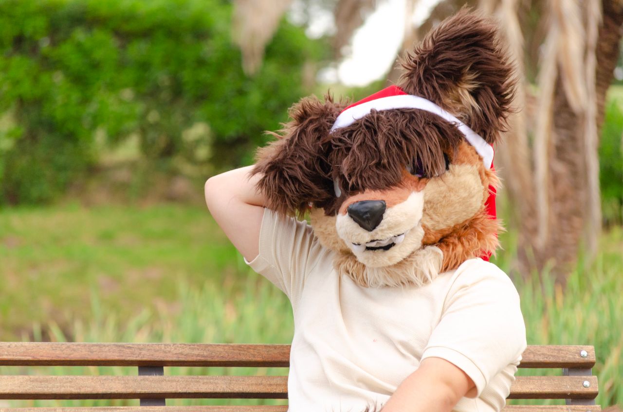 A person in a furry animal costume with a Santa hat sits relaxed on a wooden bench outdoors.